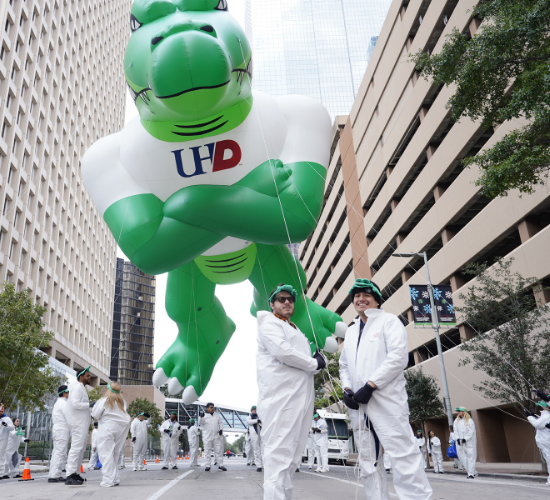 Two adults stand together at the heb thanksgiving day parade. The Ed-U-Gator parade balloon and volunteers from the university of houston-downtown stand behind them. 