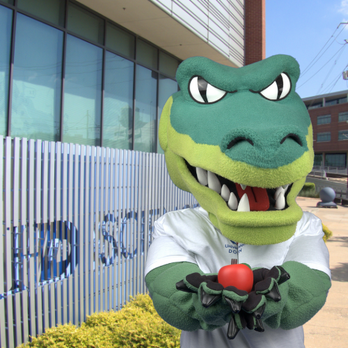 Ed-U-Gator holds an apple in front of the college of sciences and technology at the university of houston downtown