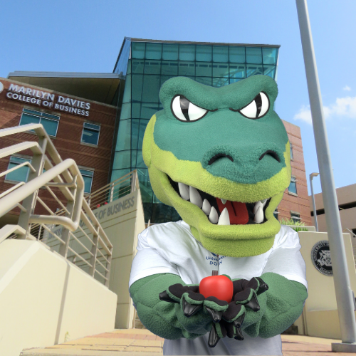 UHD mascot Ed u gator holds a red apple in front of the marilyn davies college of business building at the university of houston downtown