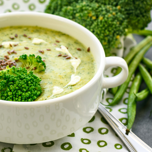 a cup of broccoli cheese soup on a table set with a spoon, broccoli and snap peas